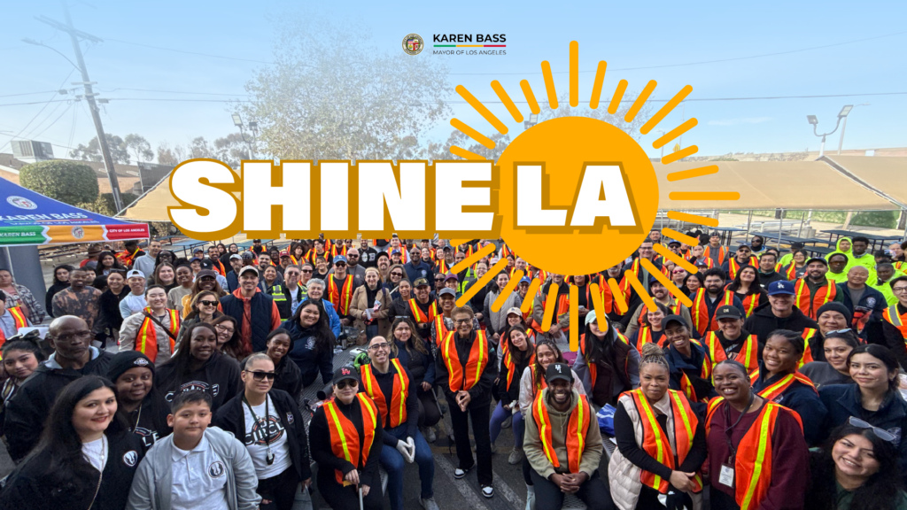 A large, diverse group of volunteers wearing orange safety vests smile and pose together for a photo at a community cleanup event. In the background, a blue City of Los Angeles canopy tent is visible. Above the crowd, bold yellow text reads “SHINE LA” with sun rays extending from the “LA” to symbolize brightness and civic pride. The image captures a vibrant, unified spirit of service and community action.